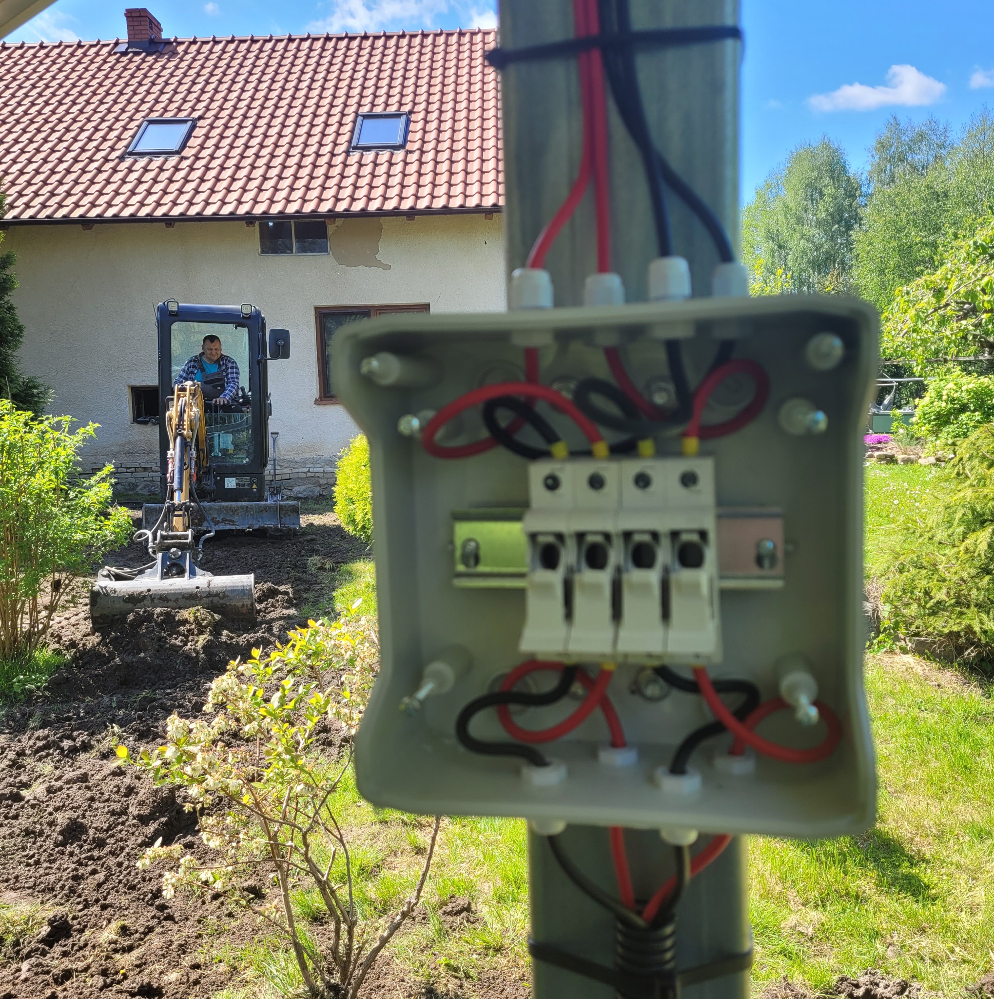 Otwarta skrzynka elektryczna z przewodami na słupie, w tle koparka przygotowująca teren pod instalację.