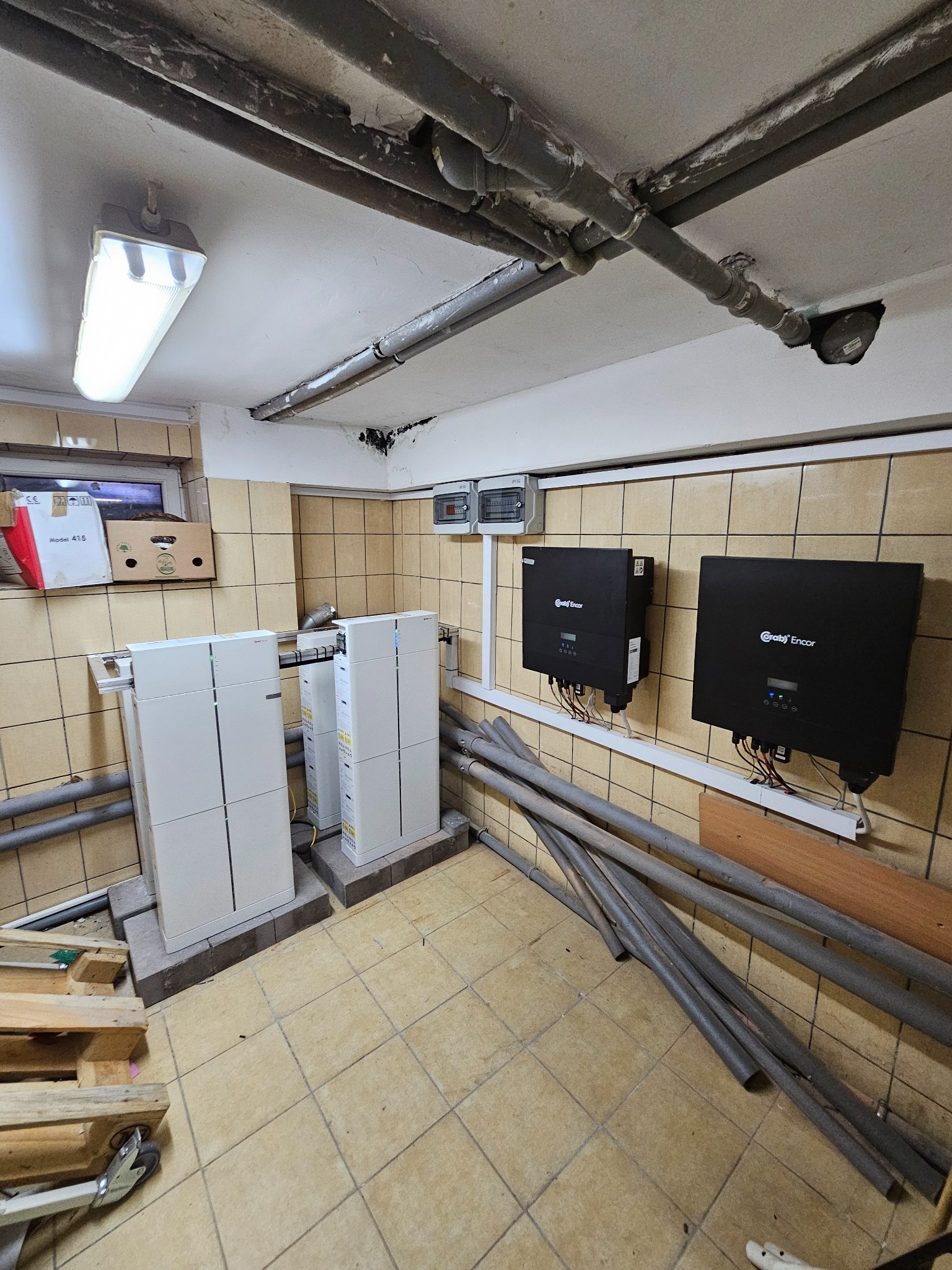 Piwniczne pomieszczenie z dwoma białymi modułami magazynu energii, dwoma czarnymi falownikami na ścianie w tle, oraz rurami i paletami na podłodze.
