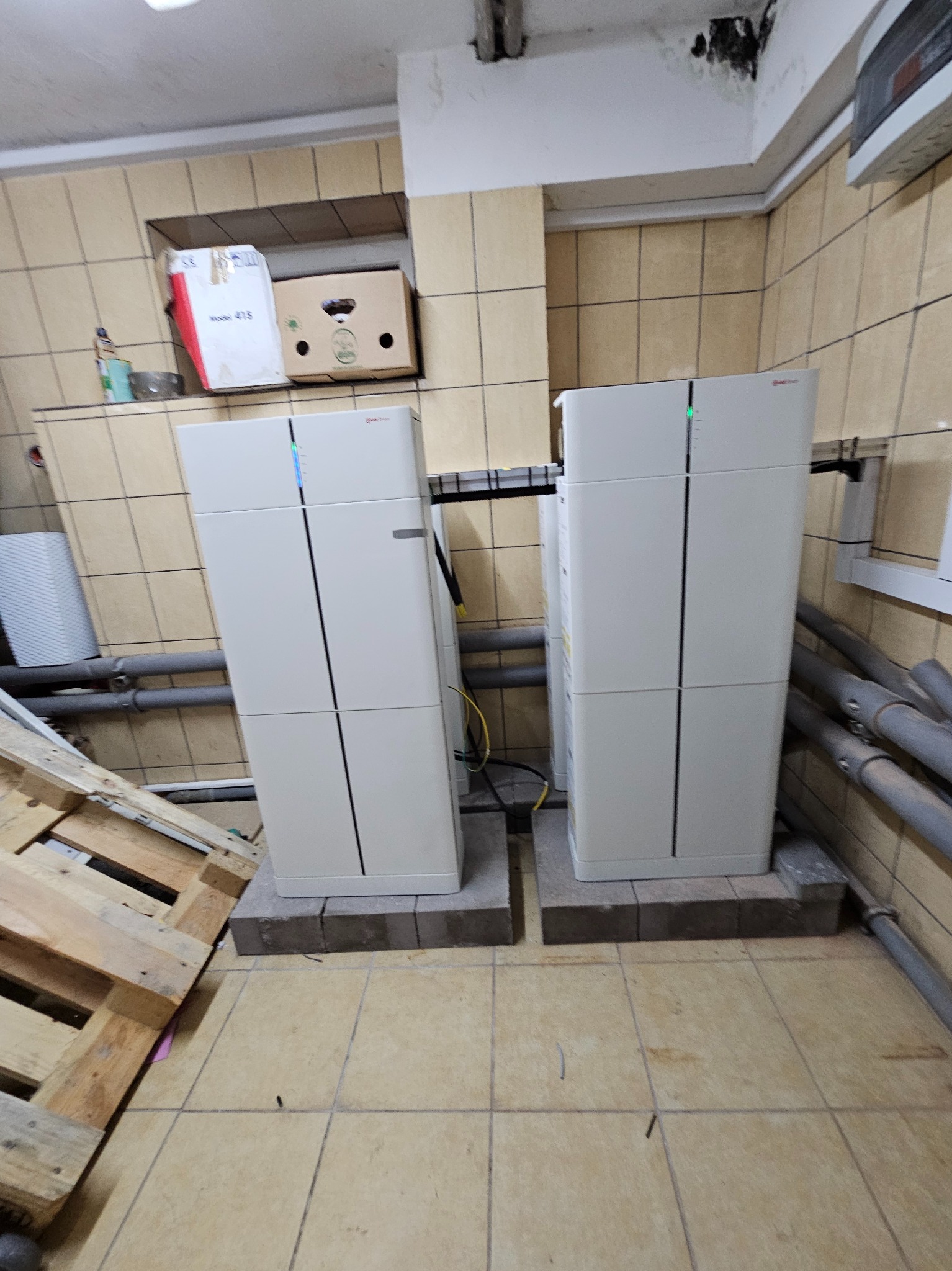 Dwa białe moduły magazynu energii na betonowych podstawach w pomieszczeniu z kafelkowymi ścianami i podłogą, widoczne rury i paleta w tle.