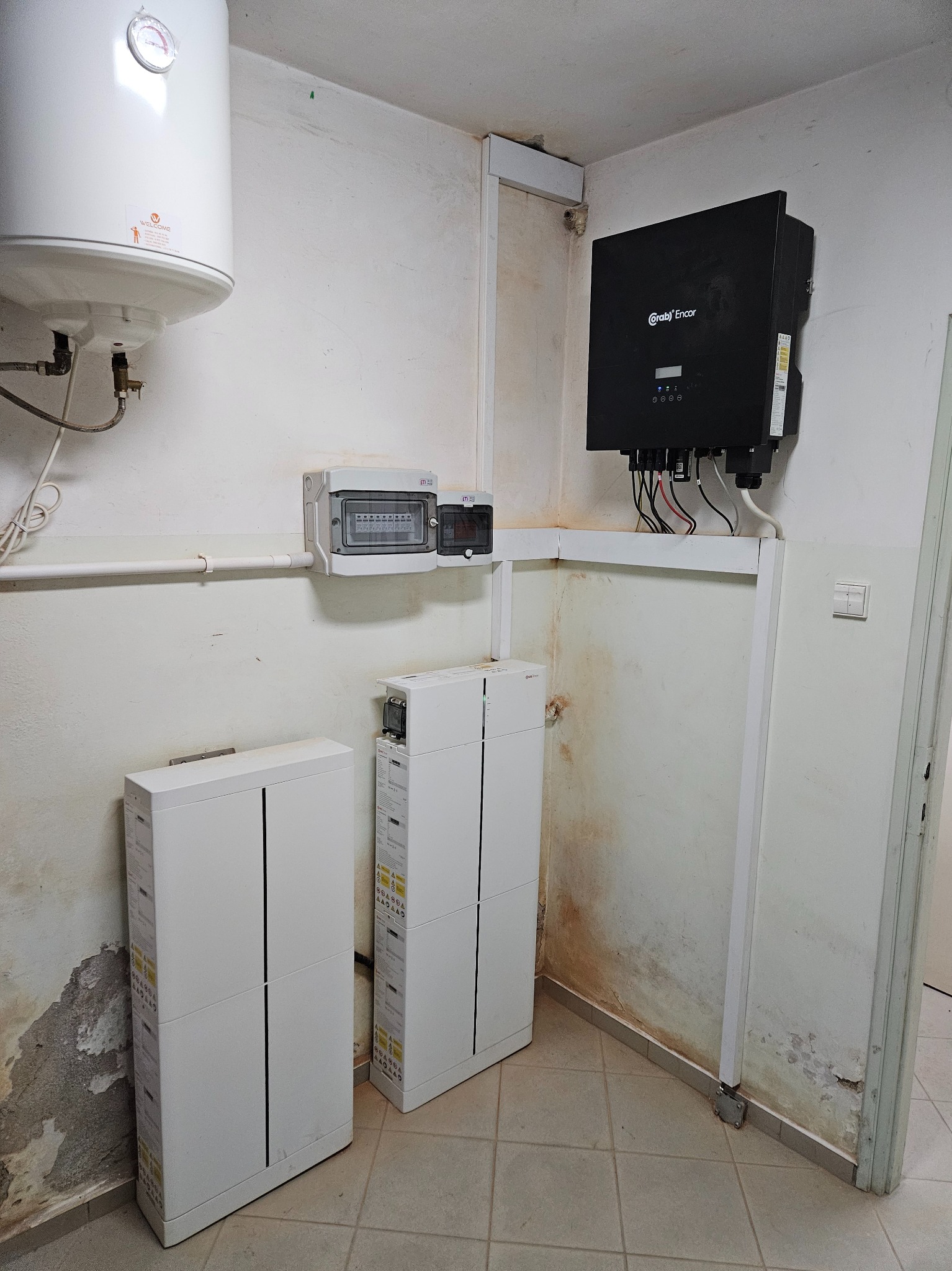 Wnętrze pomieszczenia z zamontowanym inwerterem fotowoltaicznym marki Grab'n Encor, dwoma białymi modułami magazynu energii, rozdzielnicą elektryczną i bojlerem na ścianie z widocznymi śladami...