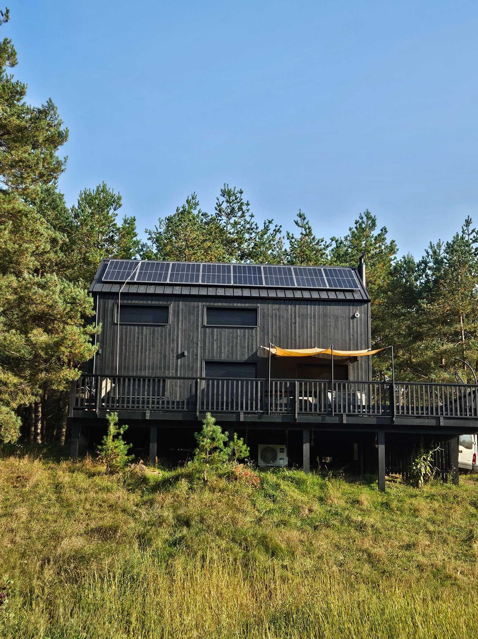 Czarny, drewniany dom na wzgórzu otoczony sosnami, z panelami słonecznymi na dachu i tarasem z żółtą markizą.