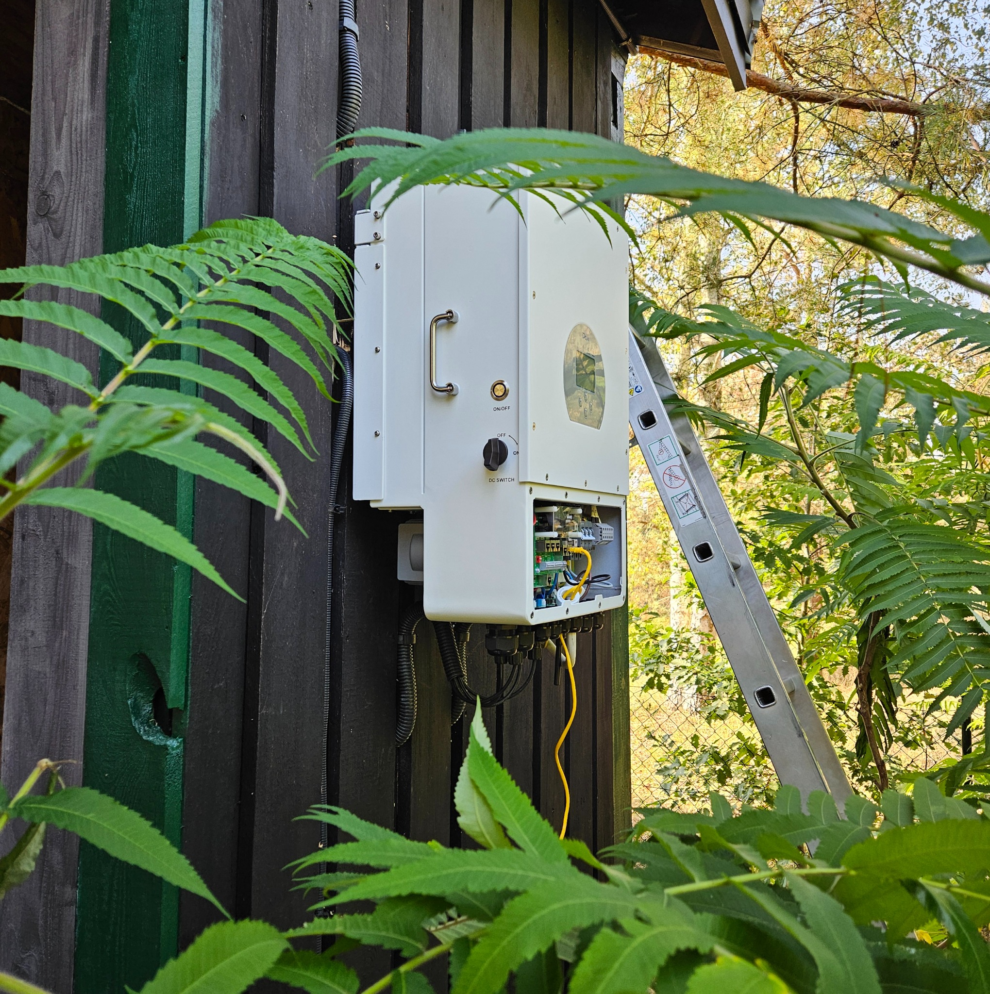 Biała skrzynka z elementami elektrycznymi zamontowana na ciemnej drewnianej ścianie budynku, z otwartą obudową ukazującą wewnętrzne okablowanie i elektronikę, obok przystawiona drabina aluminiowa...