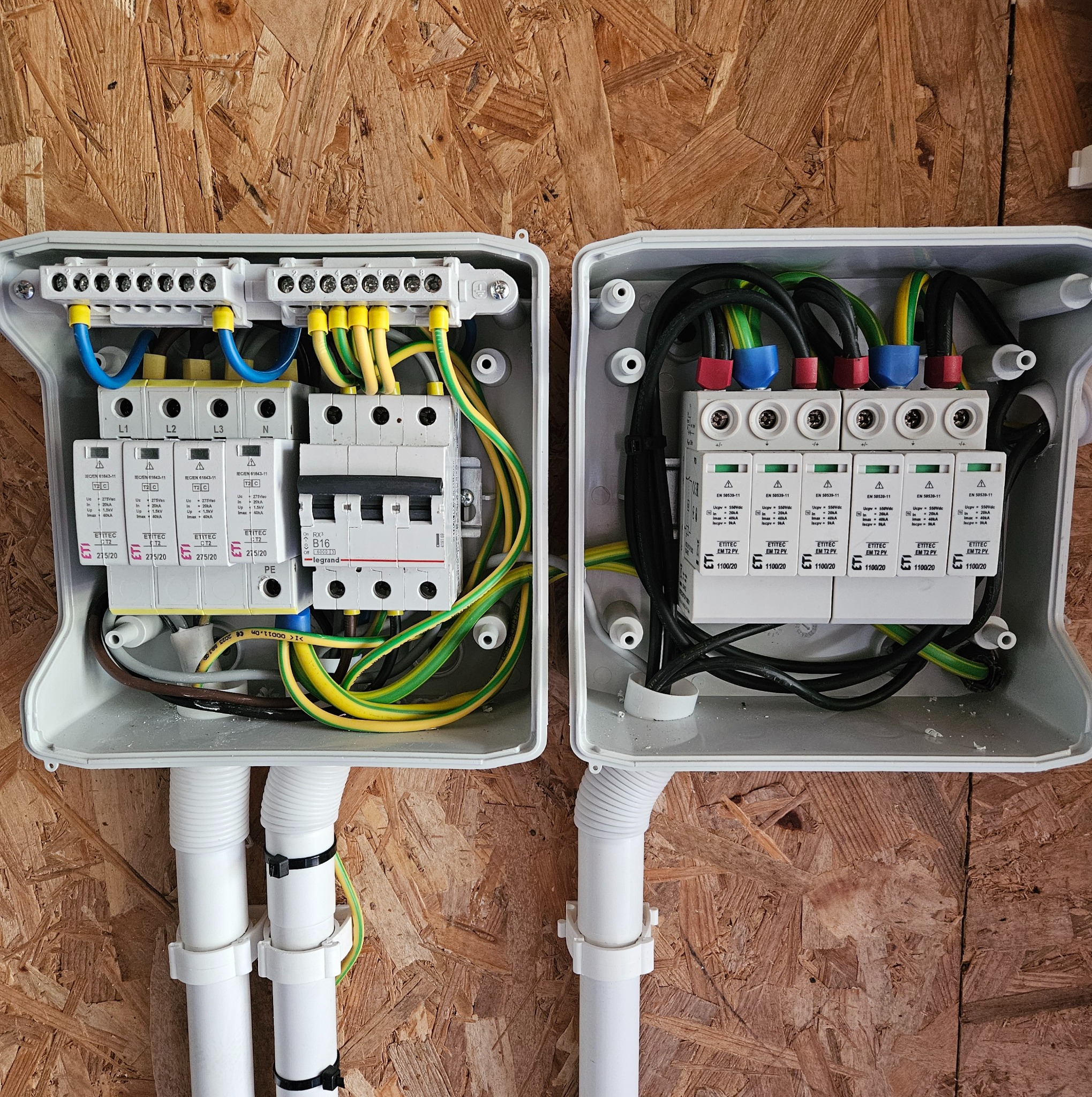 Dwa rozdzielcze panele elektryczne z zabezpieczeniami przeciwprzepięciowymi, z przewodami w kolorach żółto-zielonym, niebieskim i czarnym, zamontowane na płycie OSB.