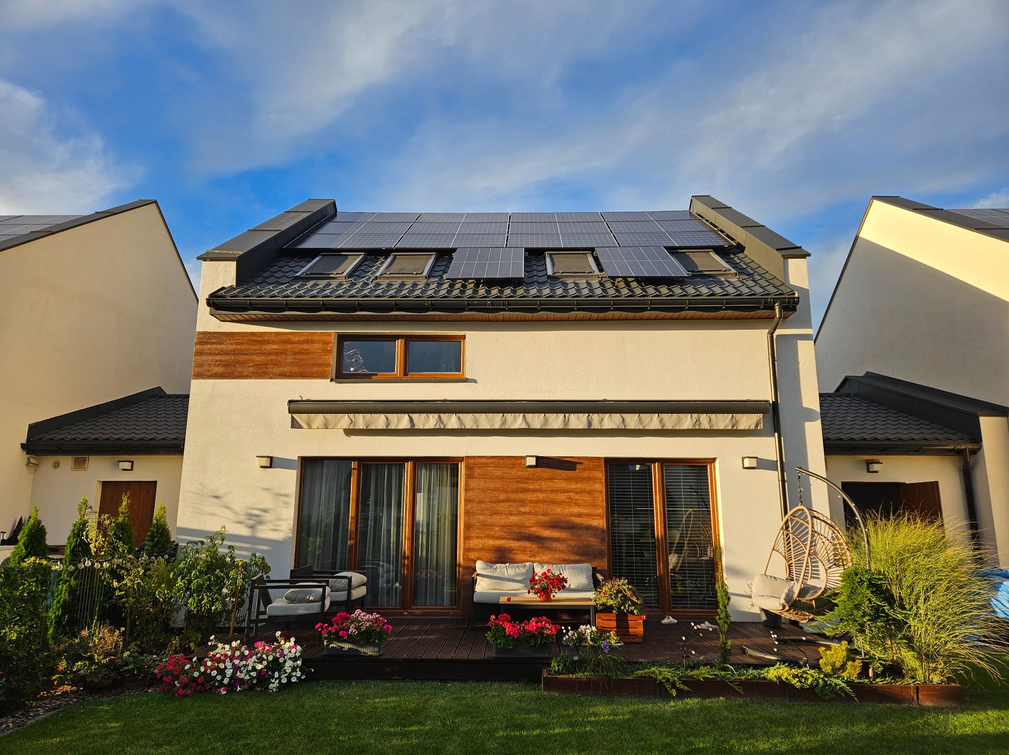 Nowoczesny dom jednorodzinny z panelami słonecznymi na dachu, widocznymi również oknami dachowymi, taras z meblami ogrodowymi i bujną zielenią w słoneczny dzień.
