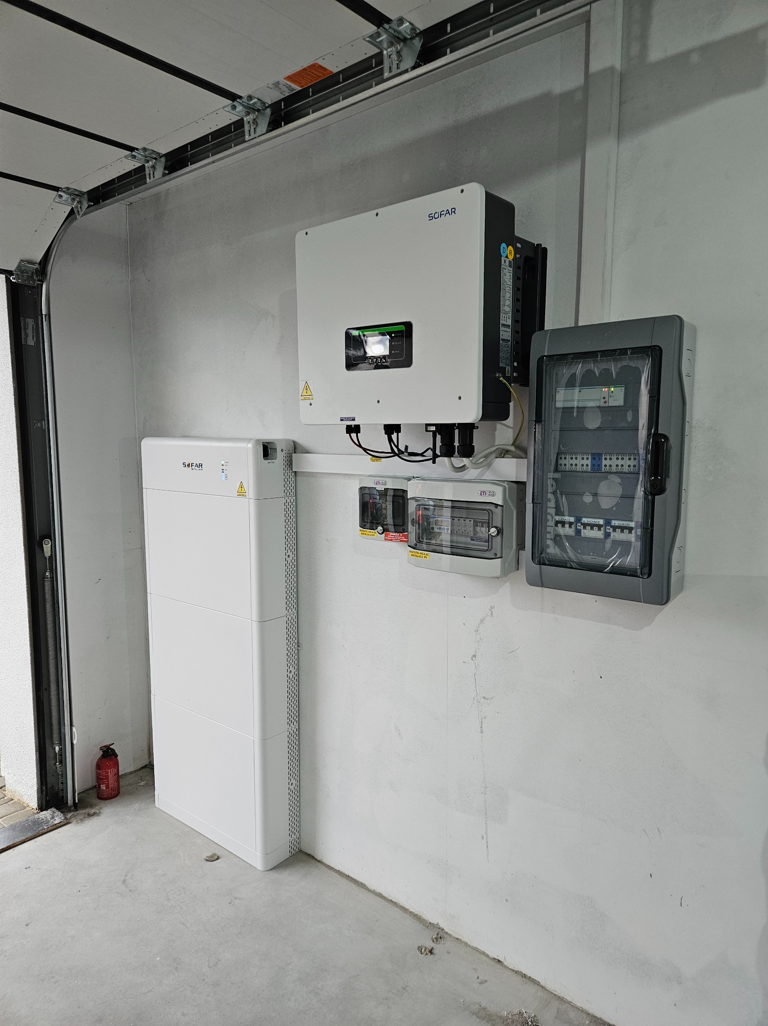 Biały magazyn energii SOFAR obok inwertera SOFAR i dwóch skrzynek z bezpiecznikami na szarej ścianie garażu, widoczna gaśnica.