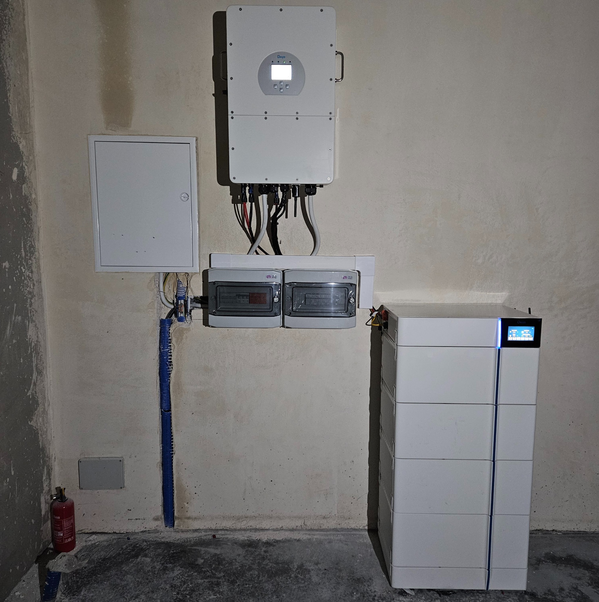 Biały magazyn energii Deye, inwerter oraz skrzynka elektryczna zamontowane na surowej, nieotynkowanej ścianie. Widoczny także niebieski peszel i gaśnica.