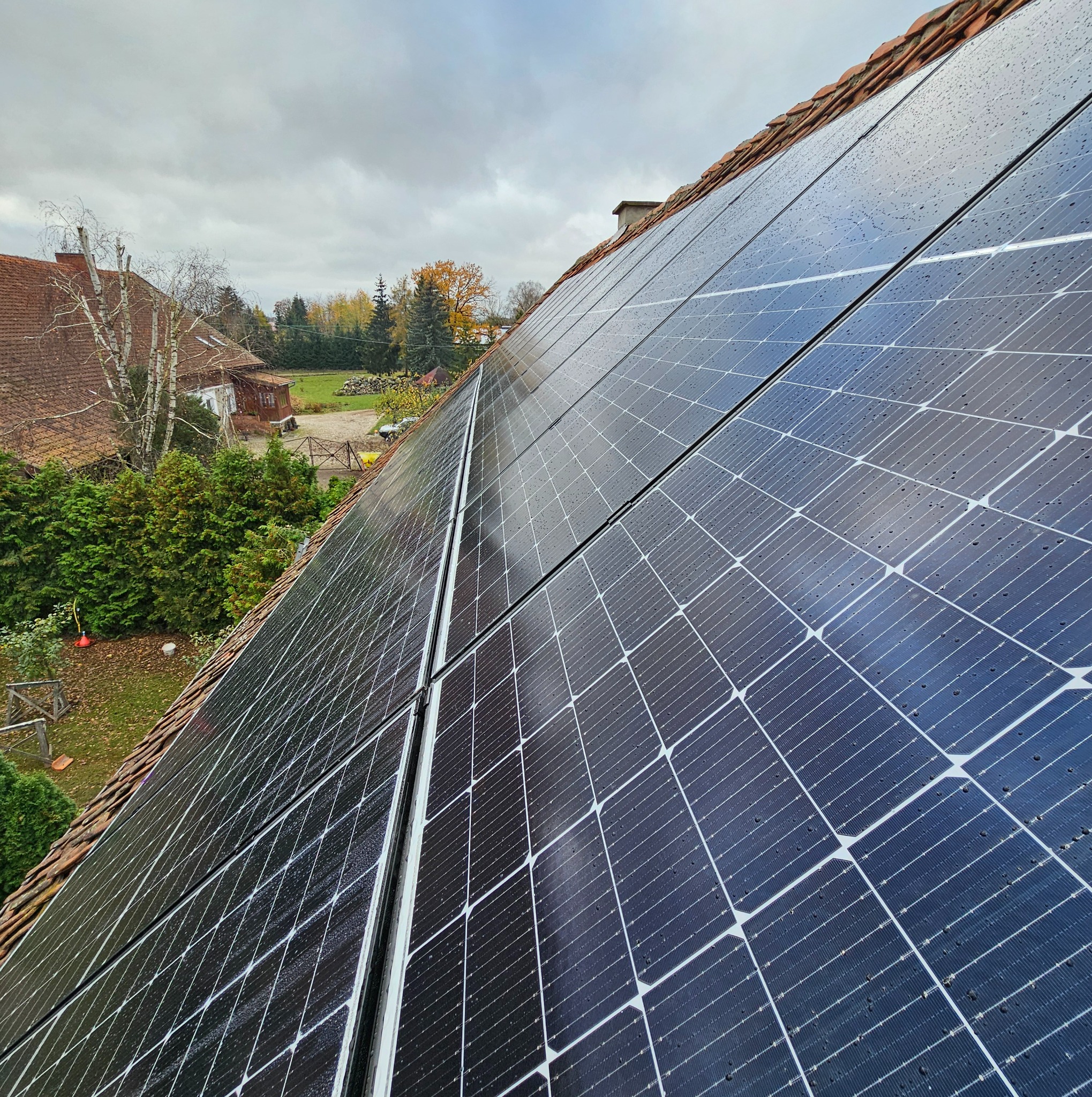 Panel fotowoltaiczny na dachu z dachówką ceramiczną, widoczne krople wody po deszczu i otoczenie domu.