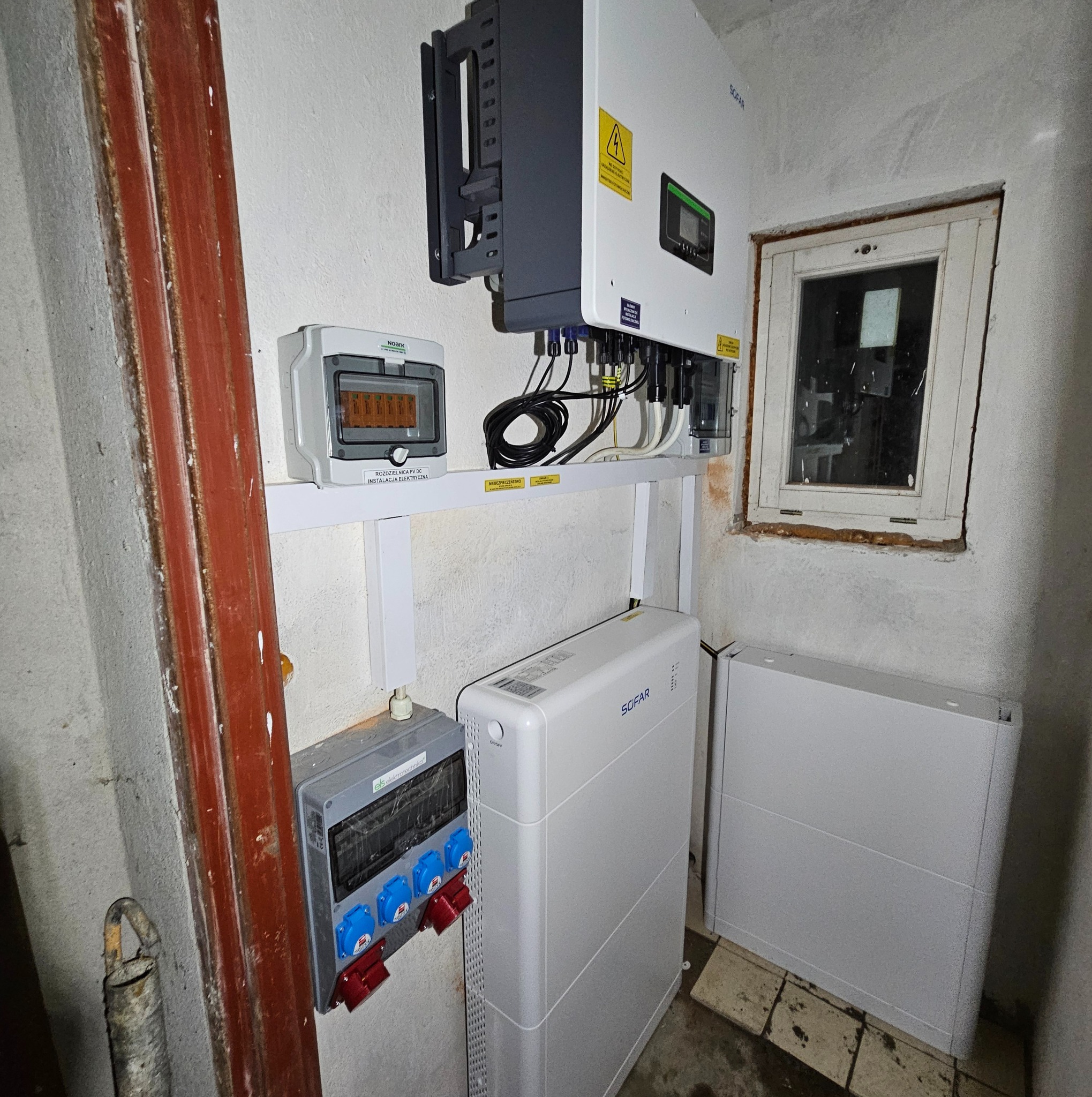 Wnętrze pomieszczenia z zainstalowanym inwerterem fotowoltaicznym Sofar, dwoma modułami magazynu energii Sofar, rozdzielnicą PV DC i gniazdami siłowymi, widoczne okno z drewnianą ramą.