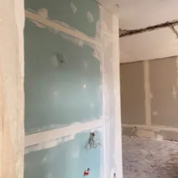 Ściana z płyty gipsowo-kartonowej w trakcie szpachlowania, widoczne wystające przewody elektryczne i niedoskonałości powierzchni przed dalszą obróbką, fragment podłogi z pozostałościami po pracach.