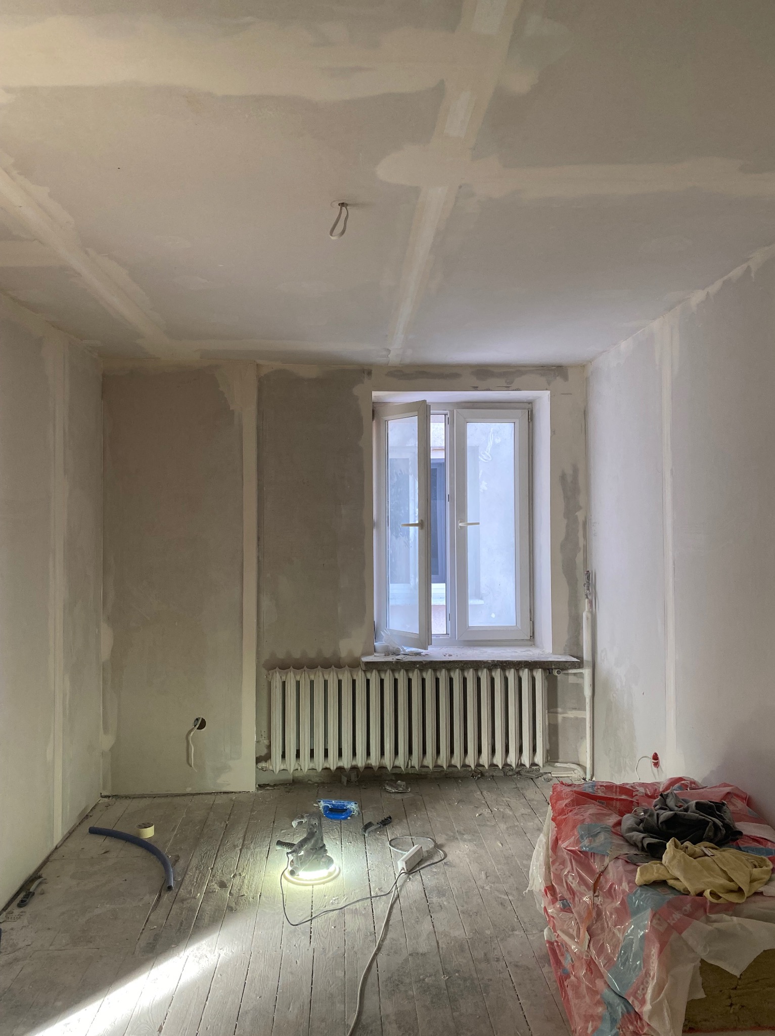 Pokój w trakcie prac wykończeniowych: ściany częściowo pokryte gładzią, szlifierka do gładzi na podłodze, okno otwarte, grzejnik żeliwny, materiały budowlane w tle.