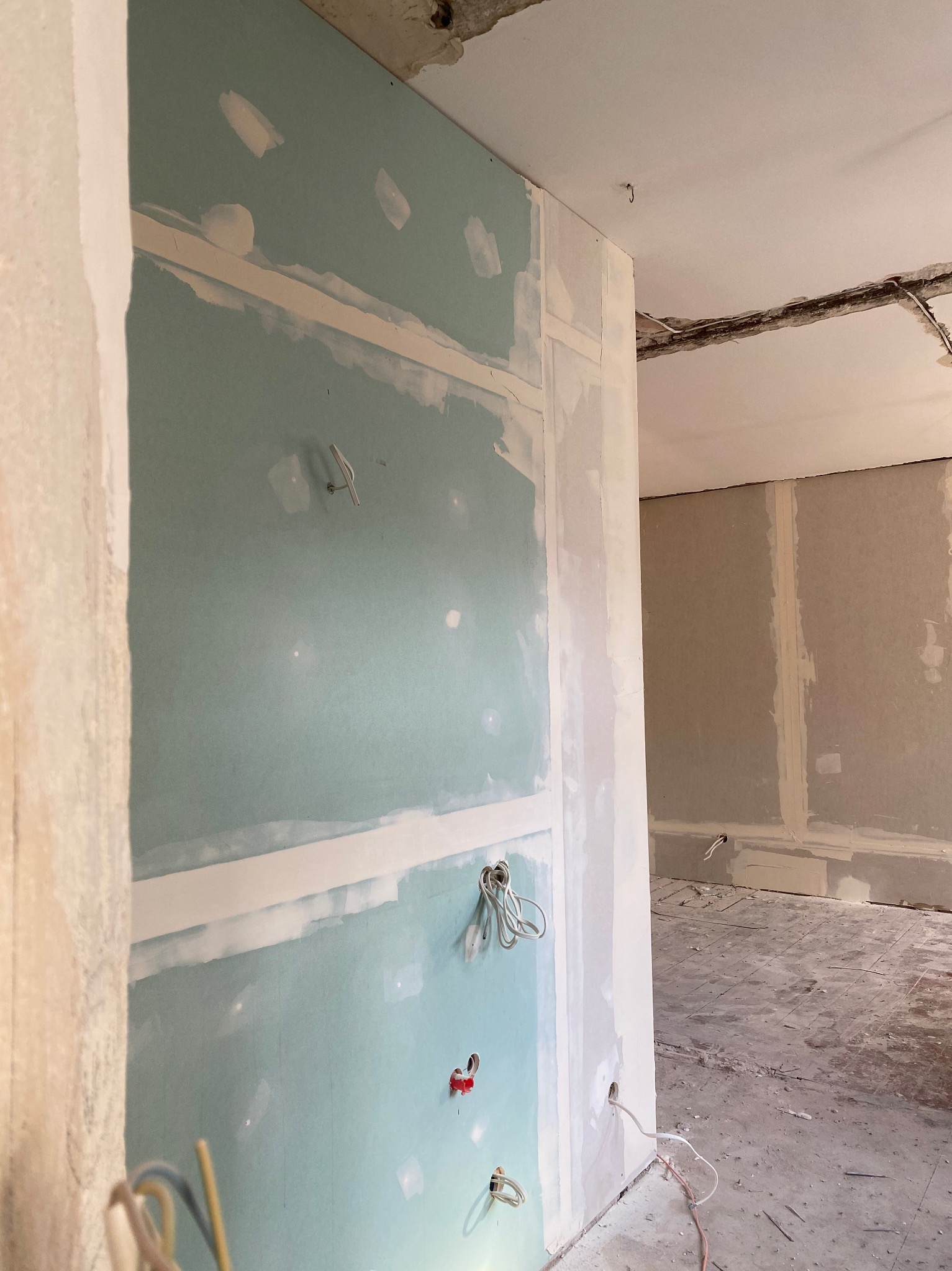 Ściana z płyty gipsowo-kartonowej w trakcie szpachlowania, widoczne wystające przewody elektryczne i niedoskonałości powierzchni przed dalszą obróbką, fragment podłogi z pozostałościami po pracach.