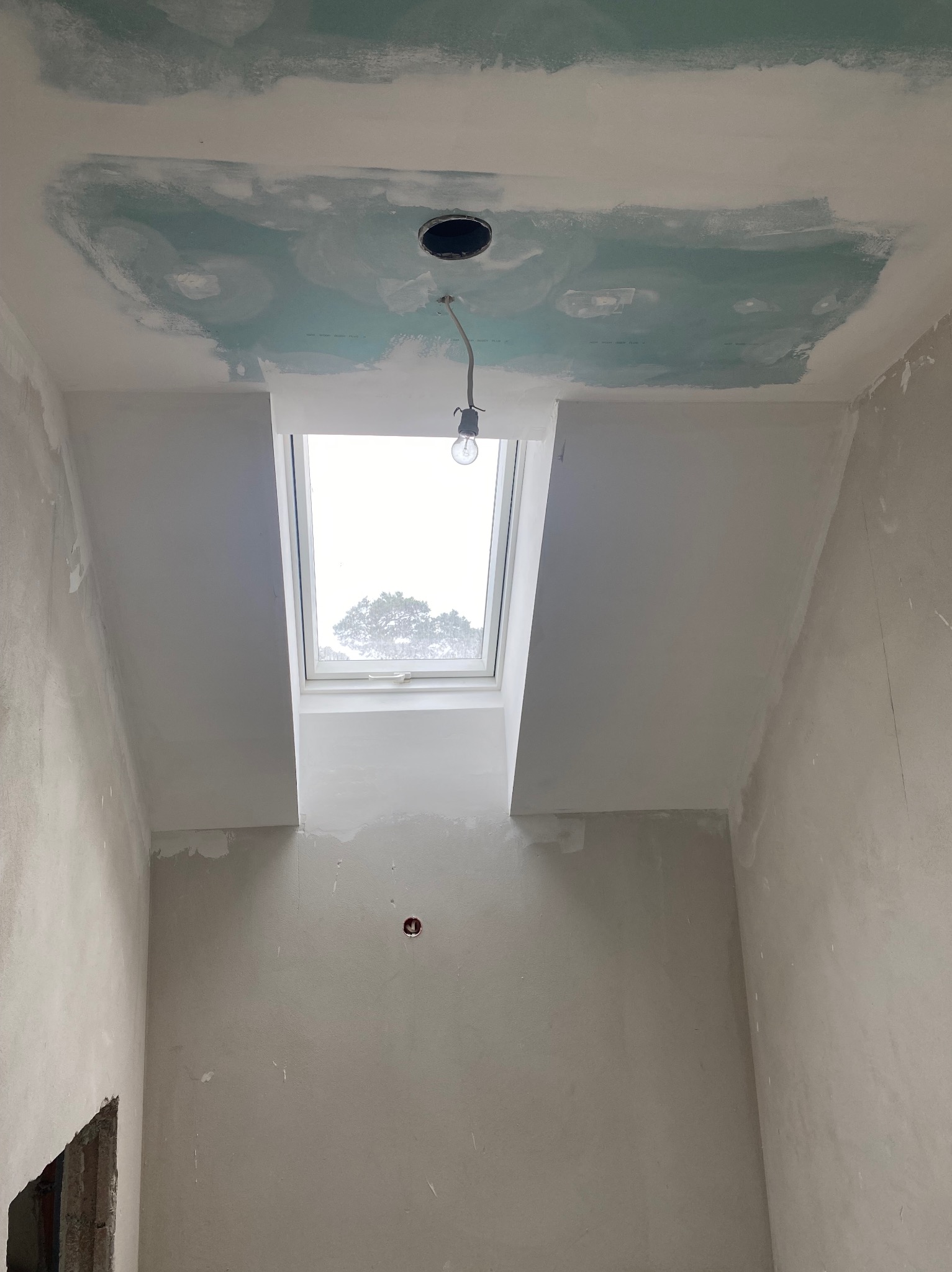 Wnętrze w trakcie prac wykończeniowych: surowa ściana, okno dachowe z widokiem na drzewo, fragment sufitu z nałożoną szpachlą i otworem na lampę z wiszącą żarówką.