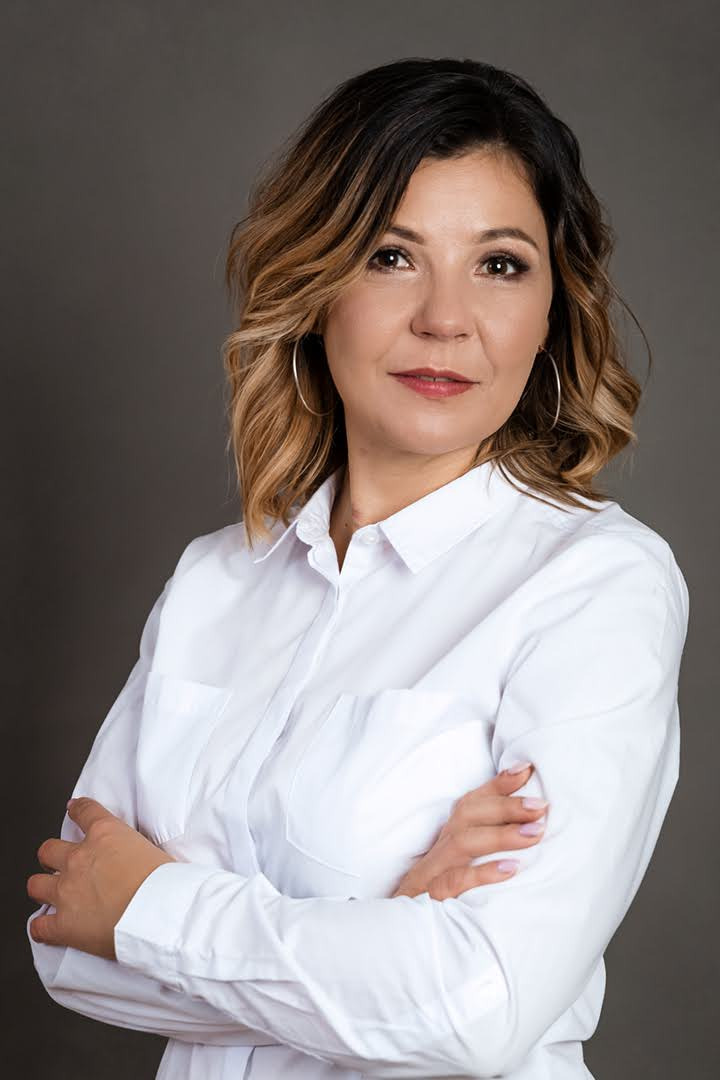 Portret kobiety w białej koszuli z założonymi rękami, z kolczykami kołami, na szarym tle.