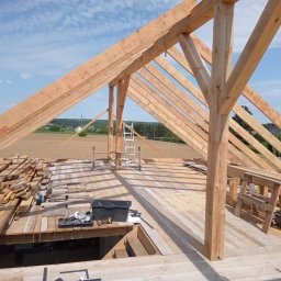 Łukasz Mazurek - Konstrukcja drewnianej więźby dachowej w trakcie montażu, widok z góry na rozłożone deski, narzędzia i krajobraz w tle.