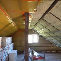 Łukasz Mazurek - Poddasze w trakcie prac izolacyjnych: widoczna folia paroizolacyjna w kolorze żółtym, wełna mineralna między krokwiami oraz zapas styropianu w opakowaniach. W tle okno połaciowe.