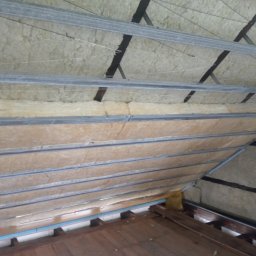 Łukasz Mazurek - Izolacja poddasza wełną mineralną, widoczne metalowe profile i fragment drewnianej podłogi, prace w toku.