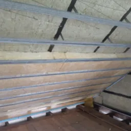 Izolacja poddasza wełną mineralną, widoczne metalowe profile i fragment drewnianej podłogi, prace w toku.