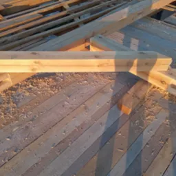 Fragment drewnianej konstrukcji dachu w trakcie budowy, widoczne elementy więźby dachowej i deski z wiórami drewna w ciepłym świetle słonecznym.