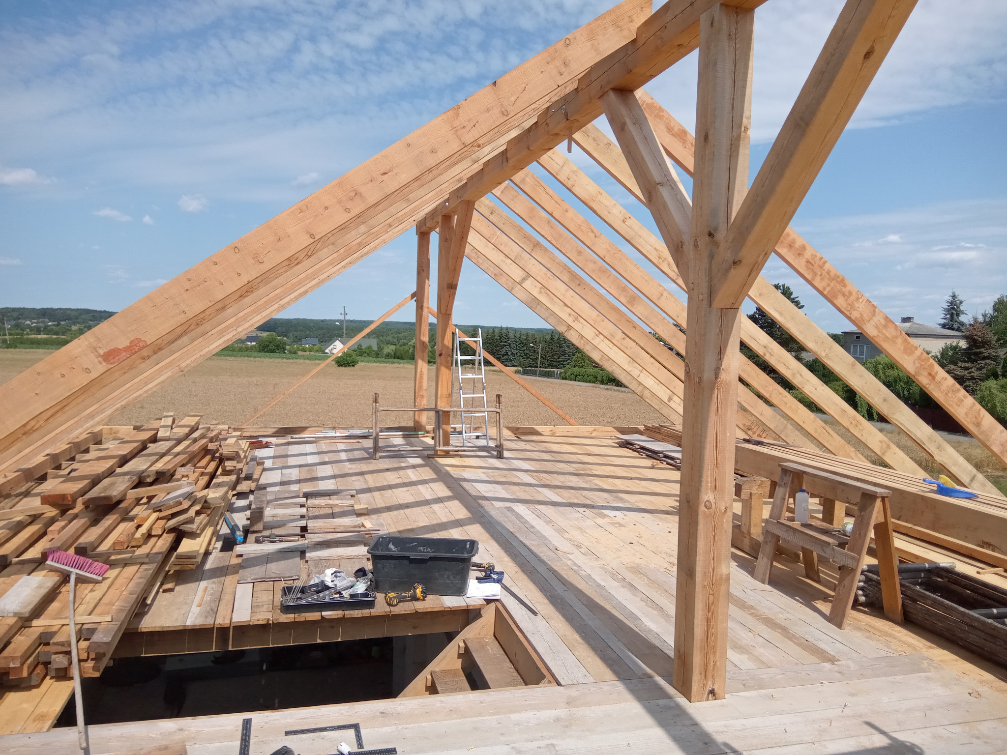 Konstrukcja drewnianej więźby dachowej w trakcie montażu, widok z góry na rozłożone deski, narzędzia i krajobraz w tle.
