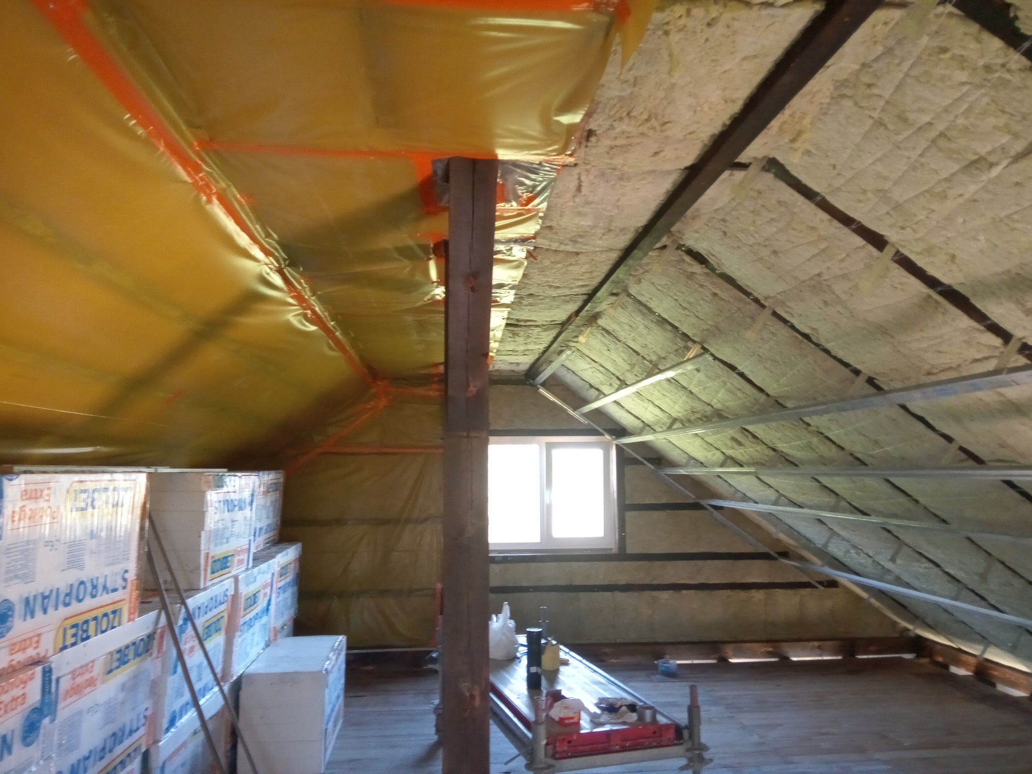Poddasze w trakcie prac izolacyjnych: widoczna folia paroizolacyjna w kolorze żółtym, wełna mineralna między krokwiami oraz zapas styropianu w opakowaniach. W tle okno połaciowe.