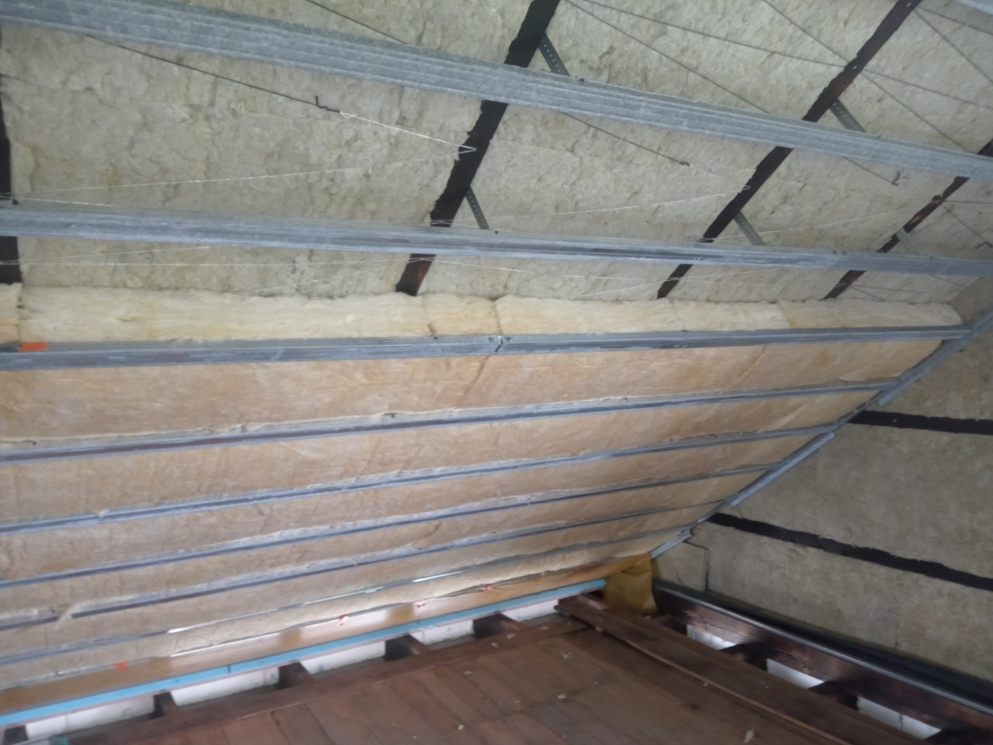 Izolacja poddasza wełną mineralną, widoczne metalowe profile i fragment drewnianej podłogi, prace w toku.