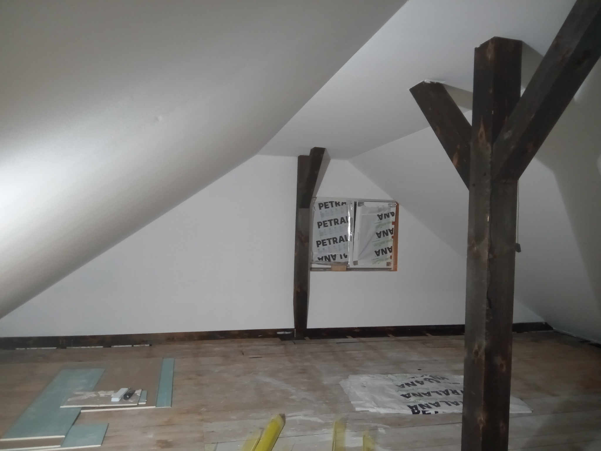 Poddasze w trakcie remontu, widoczne drewniane belki konstrukcyjne, okno zabezpieczone folią, rozłożone panele podłogowe i materiały budowlane.