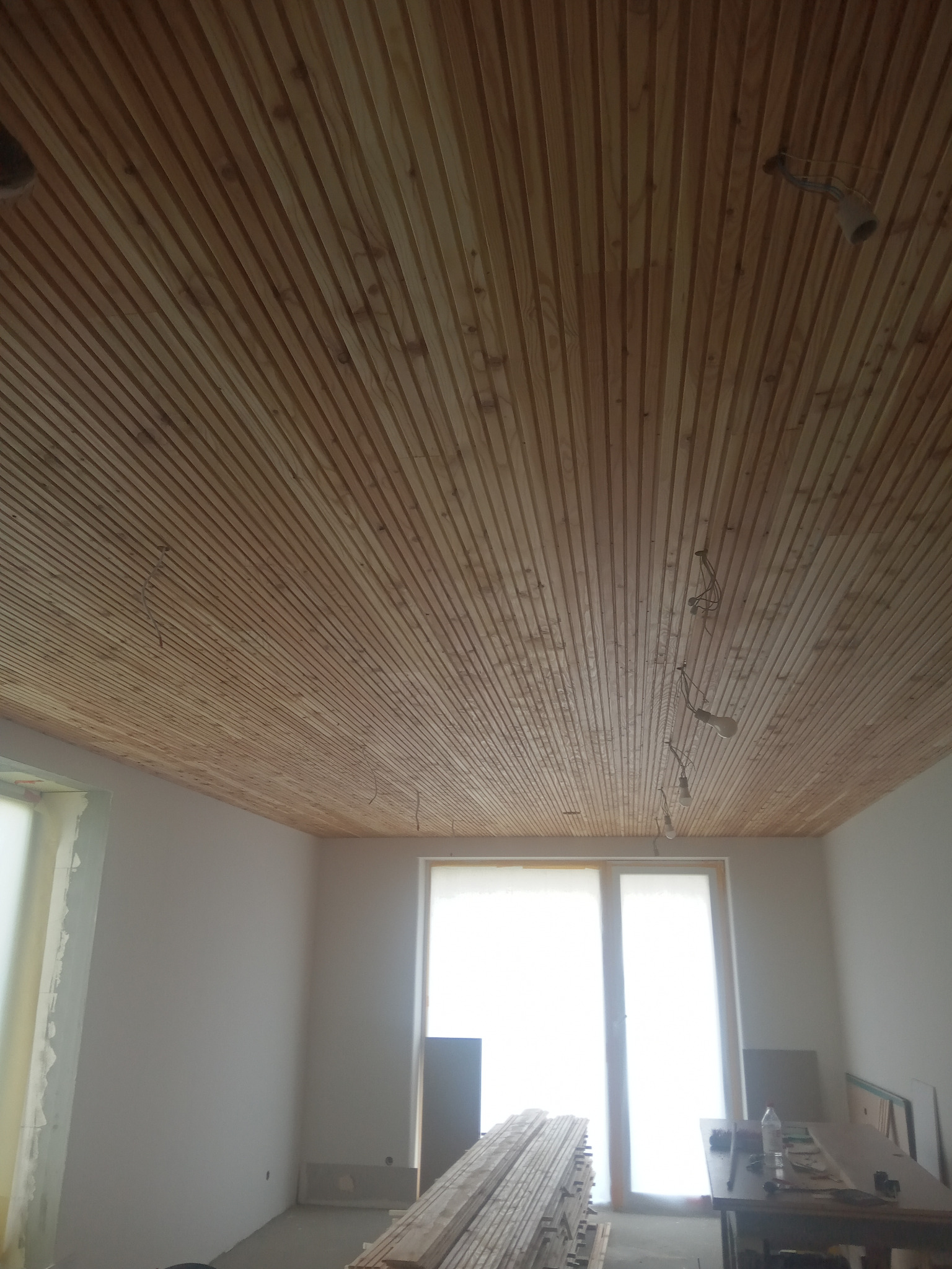 Drewniany sufit panelowy w jasnym wnętrzu, widoczne przewody oświetleniowe, okno z roletą, deski na podłodze. Ukończony montaż sufitu z boazerii.