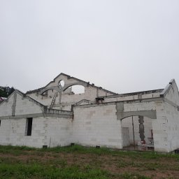 PRZEDSIĘBIORSTWO BUDOWLANO - INŻYNIERYJNE NIEDZIELA, KŁUSEK SPÓŁKA CYWILNA - Surowy stan budowy domu z białych bloczków, widoczne otwory okienne i drzwiowe, na górze konstrukcja dachu z okrągłym oknem, drabina oparta o ścianę, pochmurne niebo w tle.