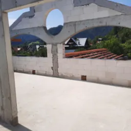 Wnętrze surowego domu w trakcie budowy, widoczne ściany z betonu komórkowego z otworem w kształcie koła i łuków, w tle krajobraz zalesionych wzgórz i zabudowań.