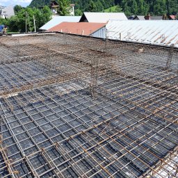 PRZEDSIĘBIORSTWO BUDOWLANO - INŻYNIERYJNE NIEDZIELA, KŁUSEK SPÓŁKA CYWILNA - Siatka z prętów zbrojeniowych ułożona na czarnej hydroizolacji, przygotowana do zalania betonem, widok z góry na tle zabudowań i zieleni.