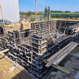 PRZEDSIĘBIORSTWO BUDOWLANO - INŻYNIERYJNE NIEDZIELA, KŁUSEK SPÓŁKA CYWILNA - Szczegółowy widok szalunków fundamentowych zbrojonych stalą, przygotowanych do zalania betonem, z widocznymi elementami konstrukcyjnymi, deskami i ziemią wokół wykopu, na tle pola i błękitnego nieba.