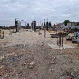 PRZEDSIĘBIORSTWO BUDOWLANO - INŻYNIERYJNE NIEDZIELA, KŁUSEK SPÓŁKA CYWILNA - Budowa fundamentów z widocznymi zbrojeniami kolumnowymi i szalunkami, teren budowy z gruzem i cegłami w tle zabudowa mieszkalna i wieża telekomunikacyjna pod pochmurnym niebem.