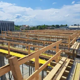 Siatka z prętów stalowych ułożona na żółtym szalunku z drewnianą barierką ochronną, widok z góry na budowę z budynkami w tle pod błękitnym niebem.