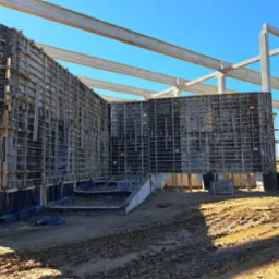 Szare szalunki fundamentowe z widocznymi elementami mocującymi, zabezpieczeniem przeciwwilgociowym i zbrojeniami w wykopie, na tle betonowych słupów konstrukcyjnych i błękitnego nieba.