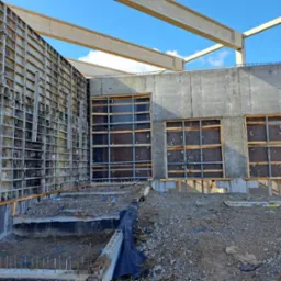 Wnętrze w surowym stanie budynku z betonowymi ścianami szalunkowymi, widoczne otwory okienne z drewnianym stelażem, fragment fundamentu z wystającymi prętami zbrojeniowymi oraz pracownik w oddali.