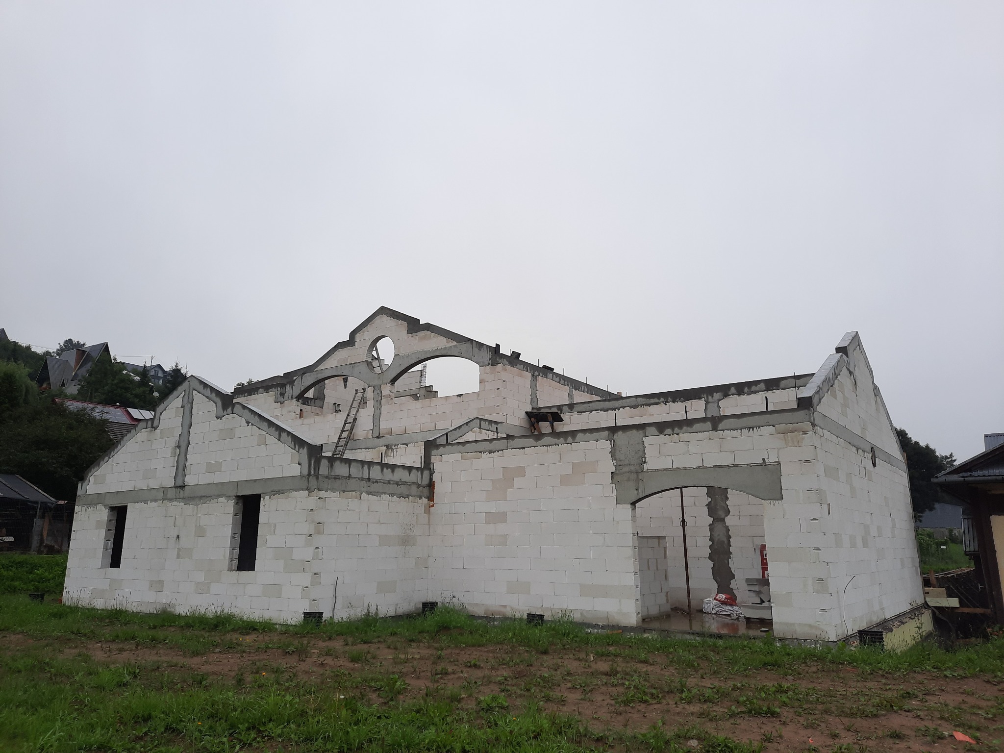 Surowy stan budowy domu z białych bloczków, widoczne otwory okienne i drzwiowe, na górze konstrukcja dachu z okrągłym oknem, drabina oparta o ścianę, pochmurne niebo w tle.