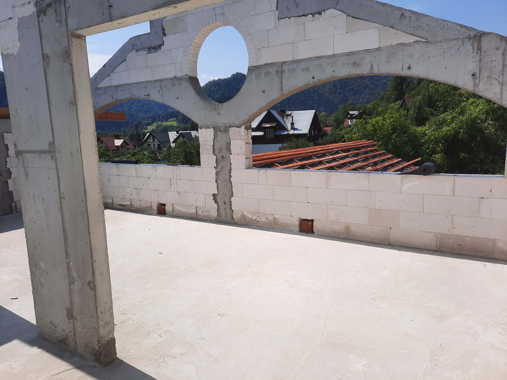 Wnętrze surowego domu w trakcie budowy, widoczne ściany z betonu komórkowego z otworem w kształcie koła i łuków, w tle krajobraz zalesionych wzgórz i zabudowań.