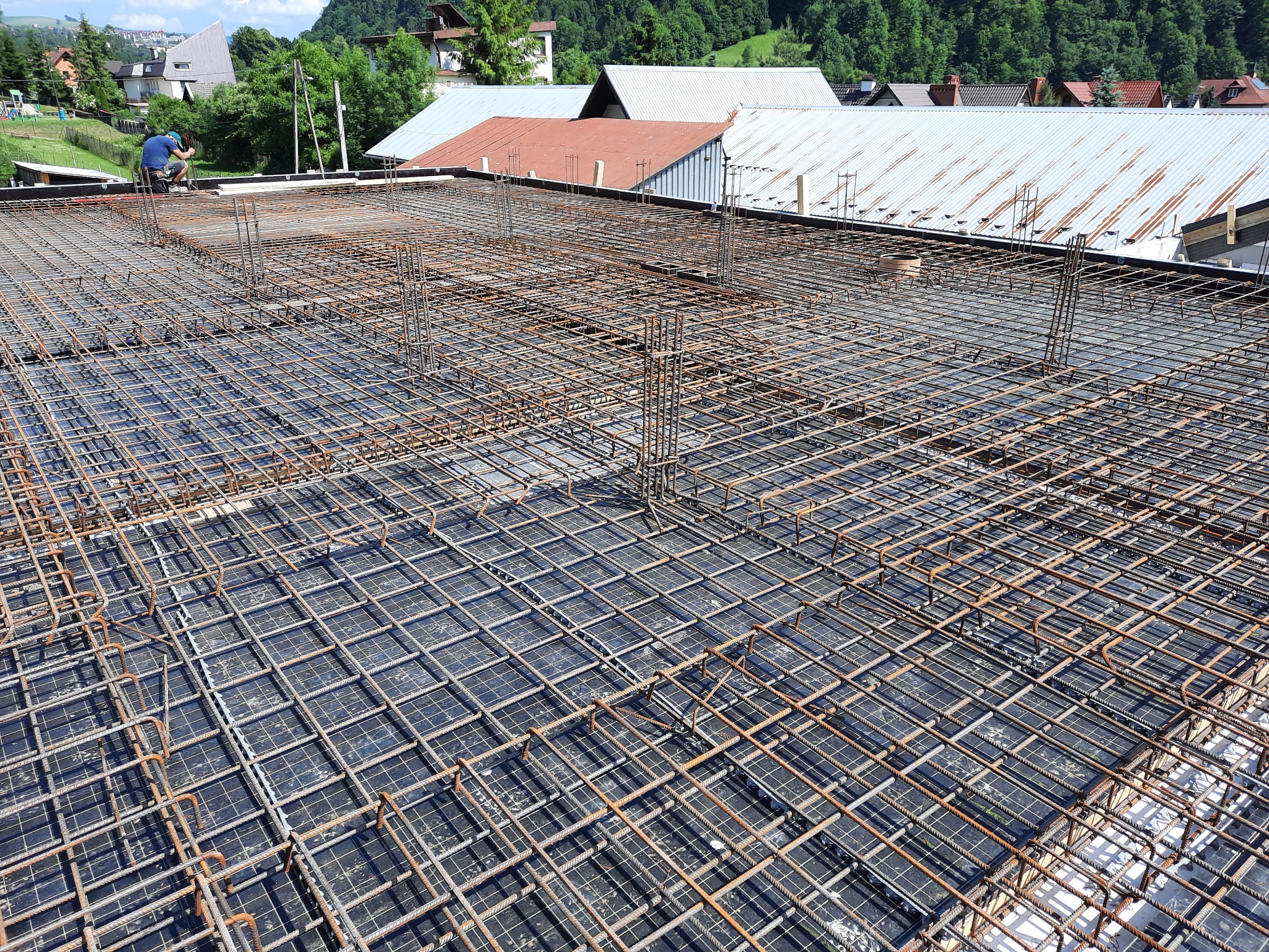 Siatka z prętów zbrojeniowych ułożona na czarnej hydroizolacji, przygotowana do zalania betonem, widok z góry na tle zabudowań i zieleni.