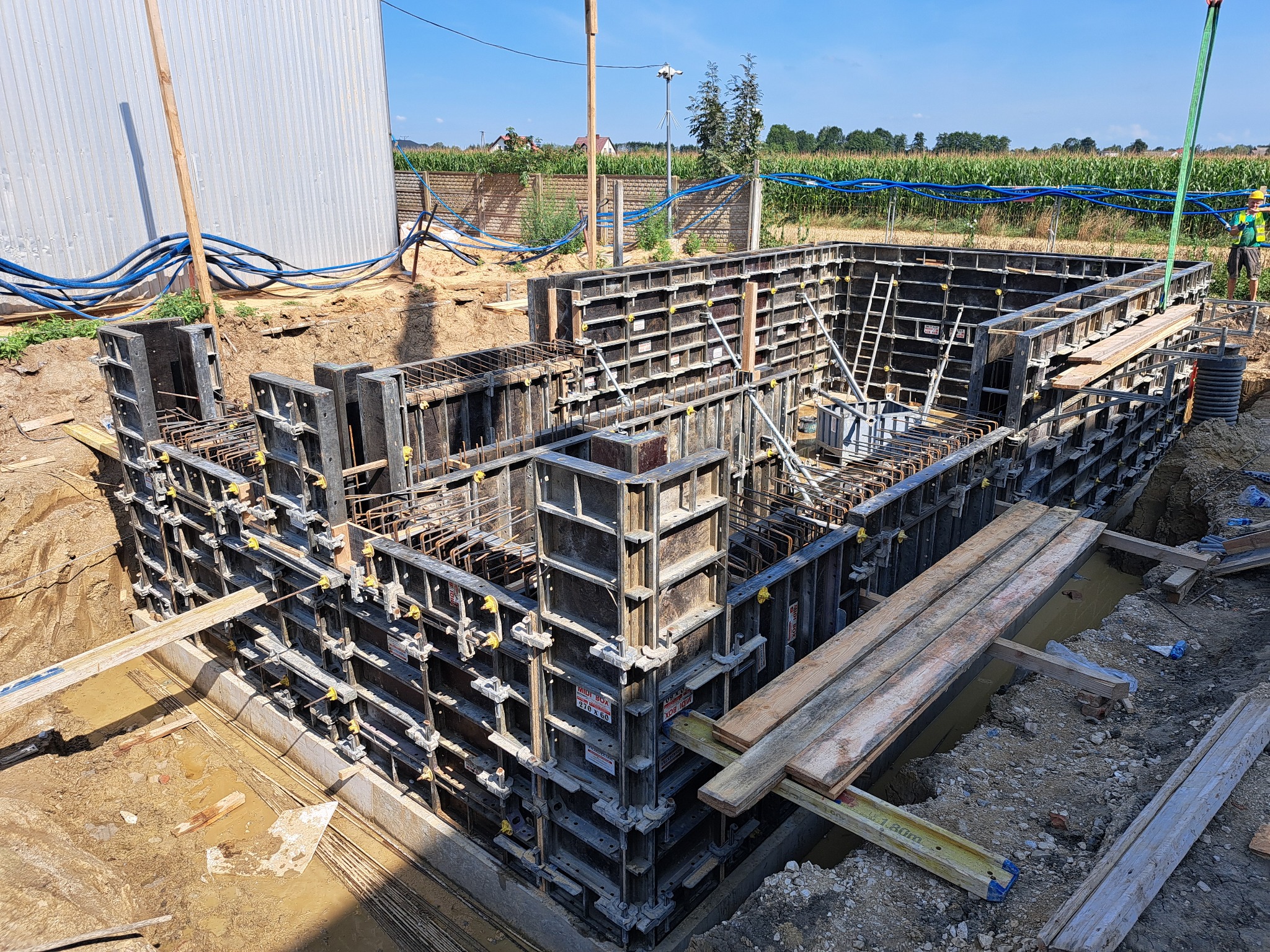 Szczegółowy widok szalunków fundamentowych zbrojonych stalą, przygotowanych do zalania betonem, z widocznymi elementami konstrukcyjnymi, deskami i ziemią wokół wykopu, na tle pola i błękitnego nieba.