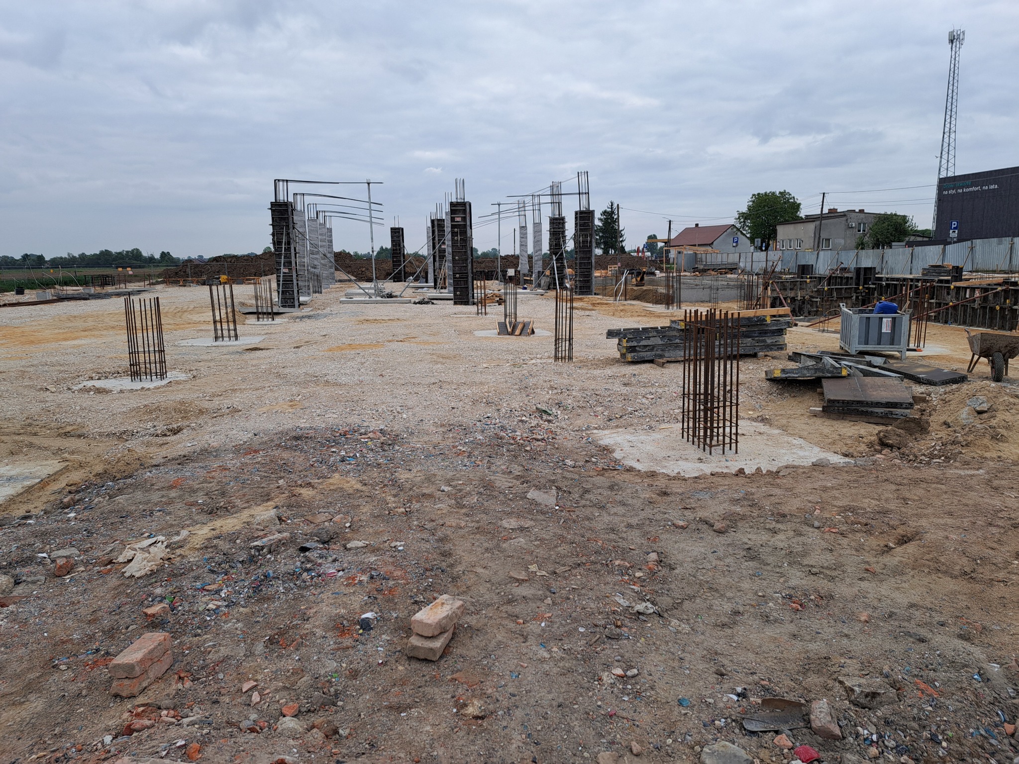 Budowa fundamentów z widocznymi zbrojeniami kolumnowymi i szalunkami, teren budowy z gruzem i cegłami w tle zabudowa mieszkalna i wieża telekomunikacyjna pod pochmurnym niebem.