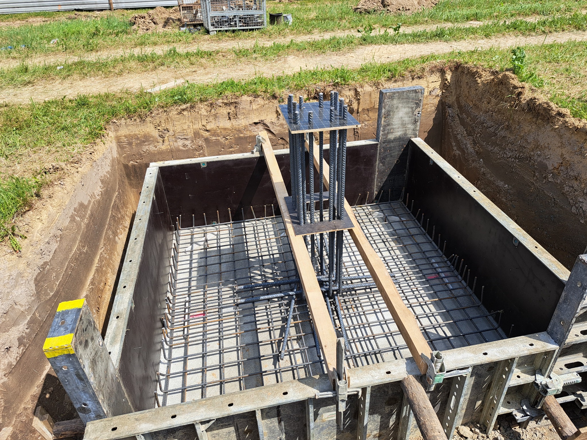 Szalunek fundamentu z siatką zbrojeniową i pionowymi prętami, zabezpieczony drewnianymi belkami, widok z góry na wykop w ziemi.