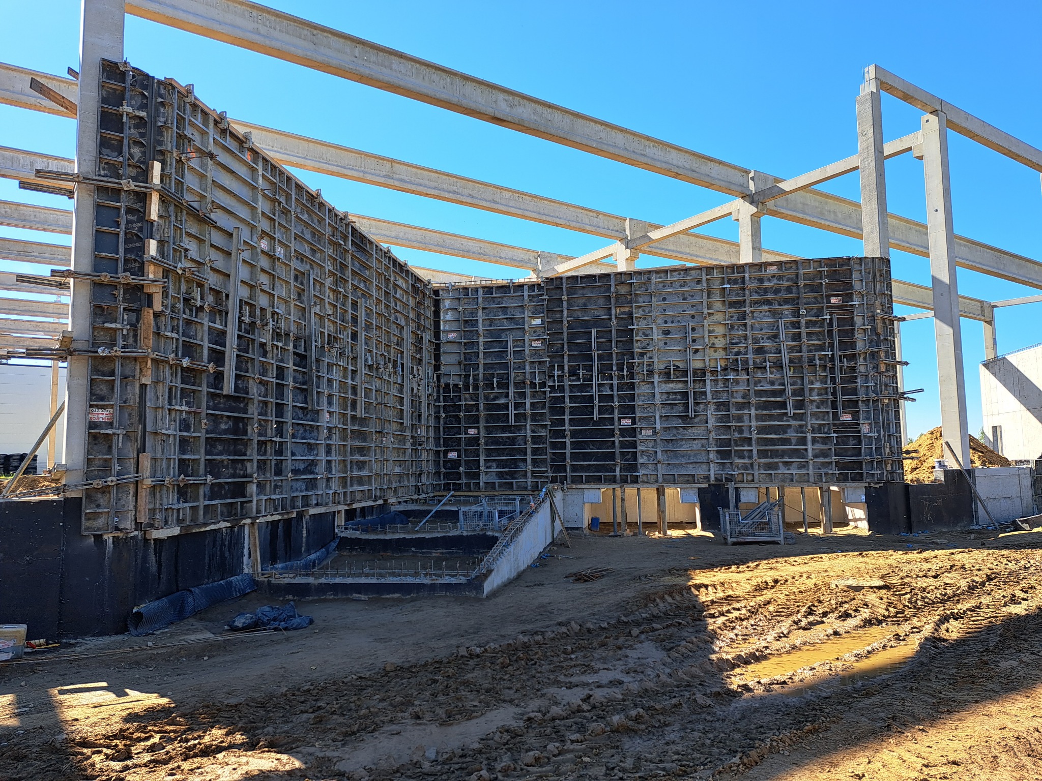 Szare szalunki fundamentowe z widocznymi elementami mocującymi, zabezpieczeniem przeciwwilgociowym i zbrojeniami w wykopie, na tle betonowych słupów konstrukcyjnych i błękitnego nieba.