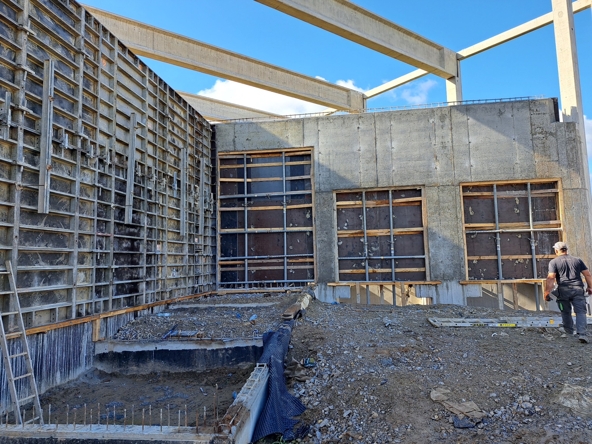 Wnętrze w surowym stanie budynku z betonowymi ścianami szalunkowymi, widoczne otwory okienne z drewnianym stelażem, fragment fundamentu z wystającymi prętami zbrojeniowymi oraz pracownik w oddali.
