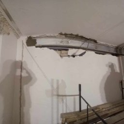 kajetankastynbud - Wnętrze w trakcie prac, z odsłoniętą konstrukcją stalową pod sufitem, fragmentami starego muru przy oknie i shadow sylwetki osoby na ścianie.