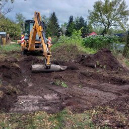 kajetankastynbud - Żółta koparko-ładowarka na działce wyrównuje teren pod budowę, widoczne ślady opon i pryzma ziemi w tle, drewniany kołek z różową farbą w lewym dolnym rogu.