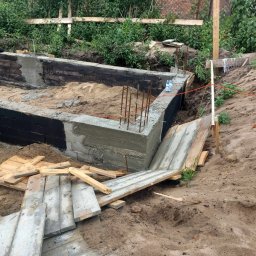 kajetankastynbud - Wykop pod budynek z częściowo wylanymi betonowymi ścianami, zabezpieczonymi czarną izolacją, z wystającymi prętami zbrojeniowymi i deskami szalunkowymi w piasku.