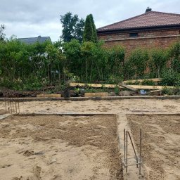 kajetankastynbud - Wykonane szalunki pod wylewkę fundamentów domu jednorodzinnego, widoczne wystające pręty zbrojeniowe, w tle budynek z czerwonej cegły i zieleń.