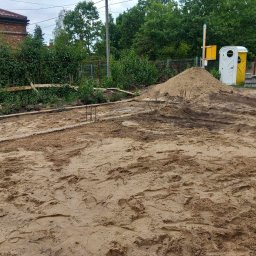 kajetankastynbud - Przygotowany teren pod budowę z widocznymi śladami stóp na piasku, drewnianym szalunkiem i wystającymi prętami zbrojeniowymi, w tle pryzma piasku, przenośna toaleta i zieleń