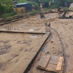 kajetankastynbud - Przygotowanie terenu pod fundamenty: wykopane rowy, zbrojenie wystające z ziemi, pracownik w tle, widoczne ślady po deszczu i błoto.