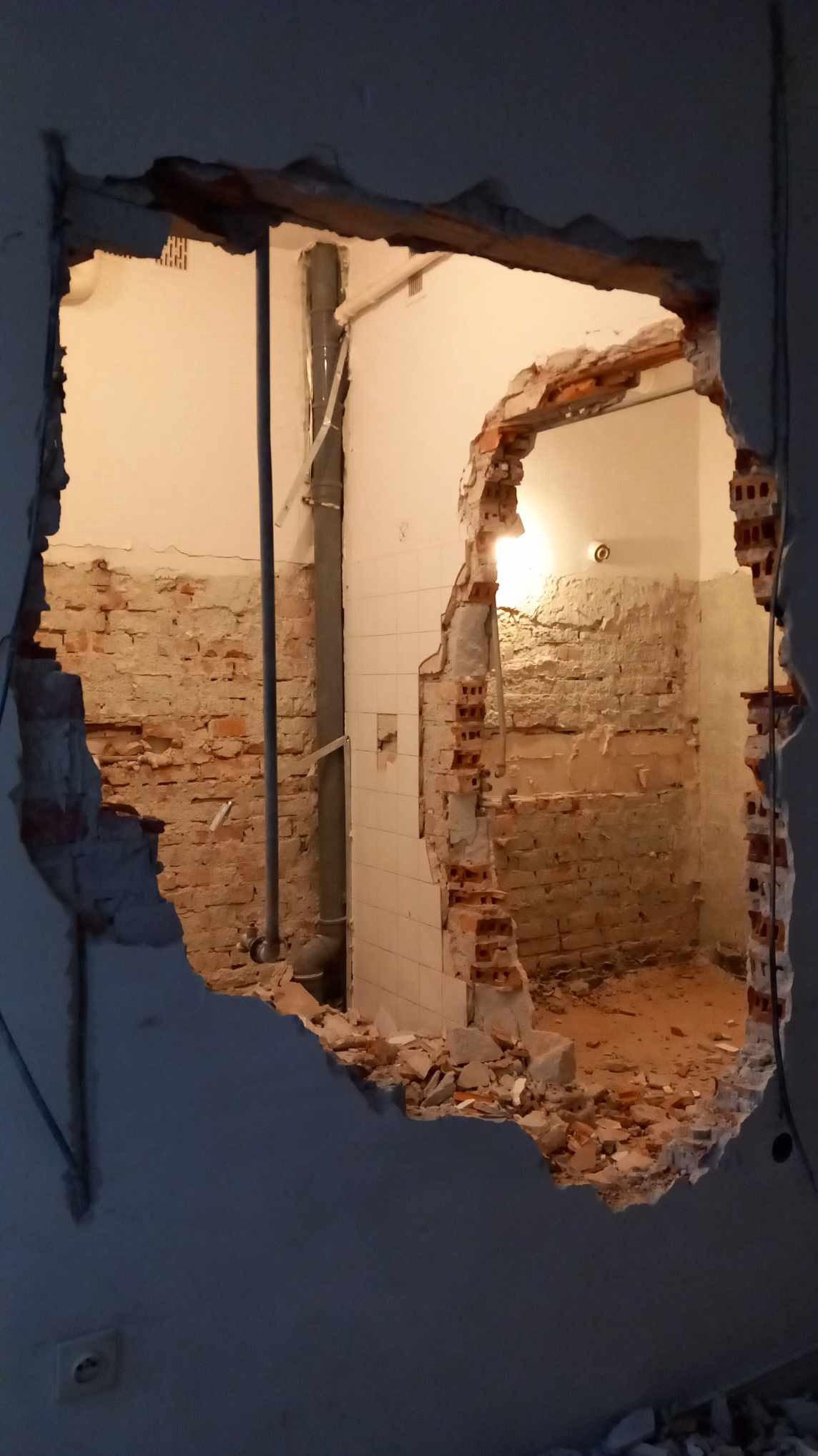 Otwór w ścianie z cegły po wyburzeniu, widoczne fragmenty instalacji i gruz, w tle fragmenty starej łazienki z białymi płytkami.
